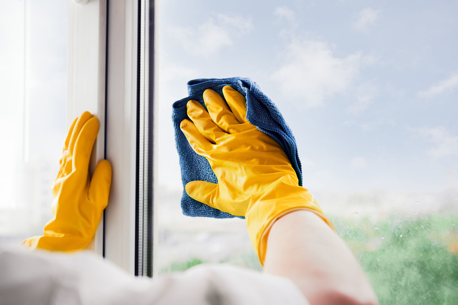 Man in yellow rubber gloves cleaning window glass with cleaner spray detergent and squeegee Housework and housekeeping, home hygiene. Blue sky view, plastic window frames.