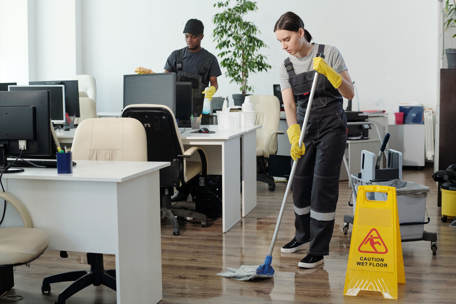 Young black man wiping computer monitors while woman with mop cleaning floor